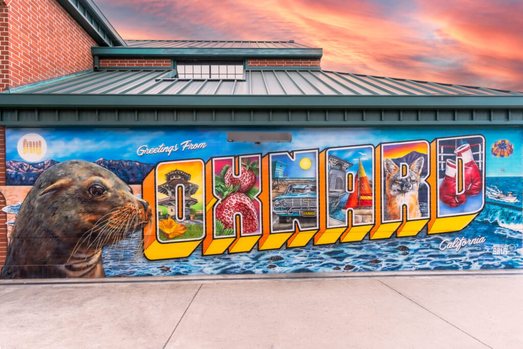 An image of a wall painted to look like a travel postcard with a realistic sea lion and the word "Oxnard" in block letters featuring famous landmarks from the city.